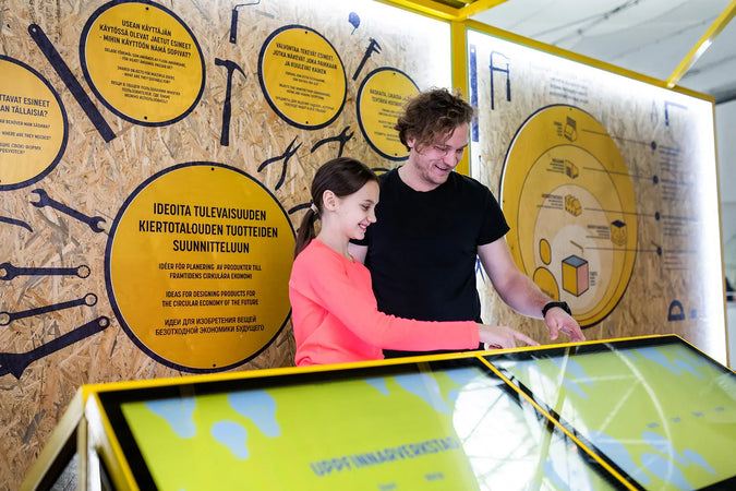 En man och en flicka provar glatt en utställning på Heurekas utställning Återvinnings Fabriken. Bakgrundsväggen är täckt med gula cirklar med information om återvinning.