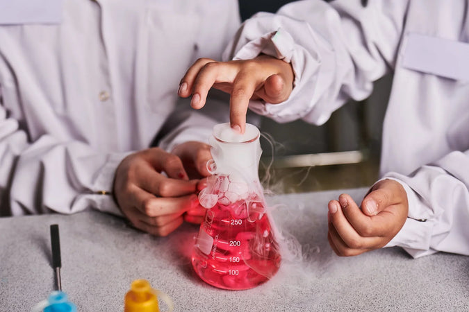 Lähikuva laboratoriotakkeihin sonnustautuneista lapsista ja heidän käsistä. Toinen lapsista kokeilee koeputken sisältöä, jossa on pinkinväristä nestettä sekä savuavaa nestemäistä typpeä.