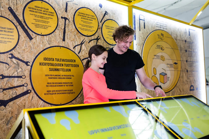 Mies ja tyttö kokeilevat iloisina Heurekan Kiertotehdas-näyttelyn kohdetta. Taustaseinällä on keltaisia ympyröitä, joissa on tietoiskuja kierrätyksestä. 