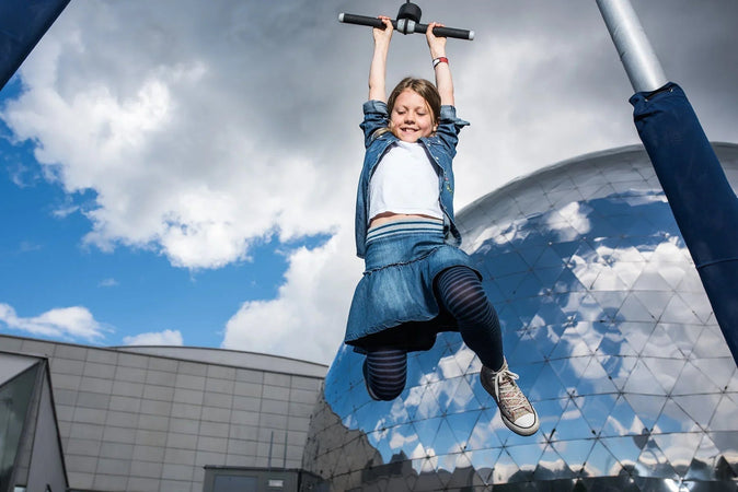 Tyttö hyppäämässä Heurekan Tiedepuisto Galilein Ihmisjojossa. Planetaarion kuvusta heijastuu sininen taivas valkoisine pilvineen.