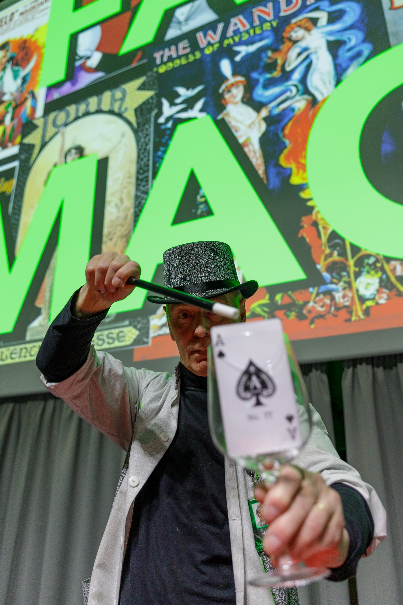 Mies pitää kädessään lasia, jossa on Ässä-kortti. Toisessa kädellä hän osoittaa lasia taikasaualla.