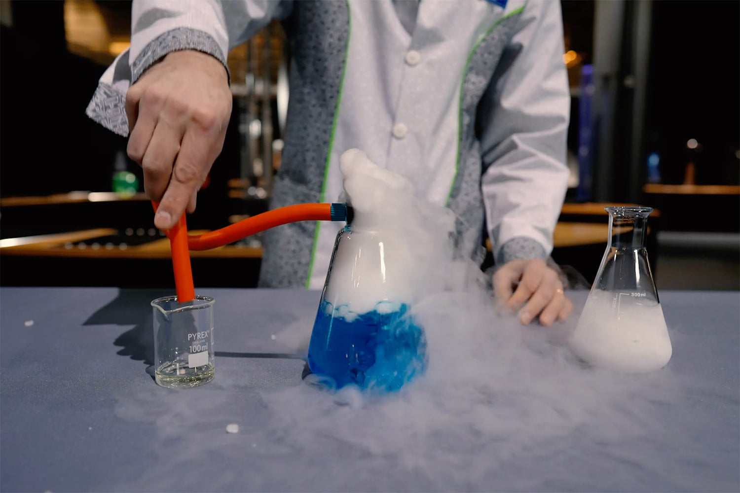 Kuvassa näkyy laboratoriotakkisen henkilön kädet ja kolme erilaista koepulloa. Yhdessä on sinistä nestettä ja höyryävää savua. Tekeillä on tiedetemppu.