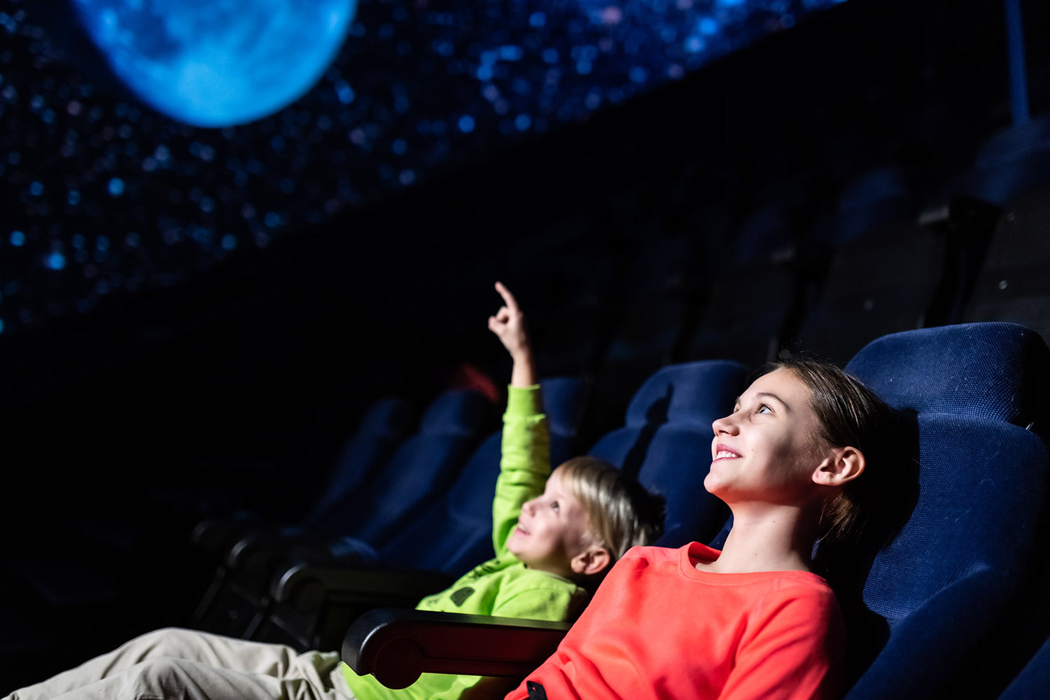 Two children are sitting in the Heureka planetarium, one of them pointing at a picture showing stars and a large moon.